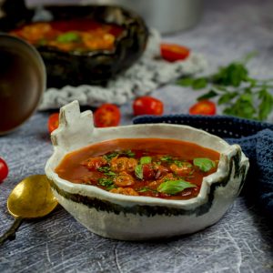 Bowl of minestrone soup on a grey table with tomatoes and herbs around it