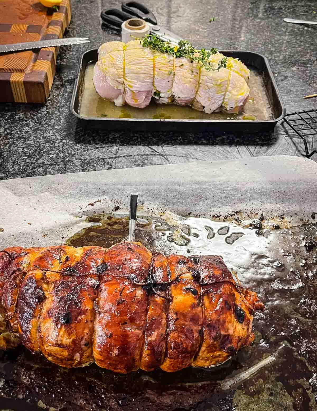 A cooked rolled roast in the foreground and an uncooked rolled roast in the background on a dark kitchen bench