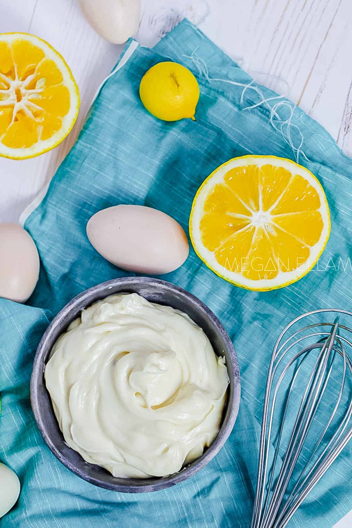 A grey bowl with mayo in it on a piece of blue fabric with eggs and lemons too.