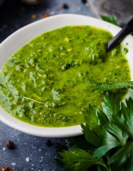 A vibrant green herby dressing in a white serving dish on a dark counter top strewn with spices and fresh herbs