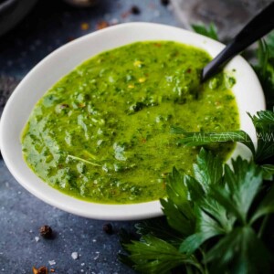 A vibrant green herby dressing in a white serving dish on a dark counter top strewn with spices and fresh herbs