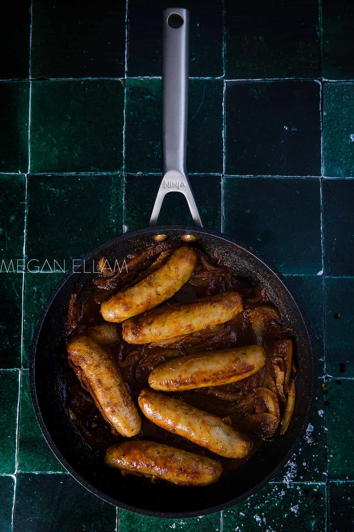 Sausages in gravy in a skillet on a green tiled backdrop.