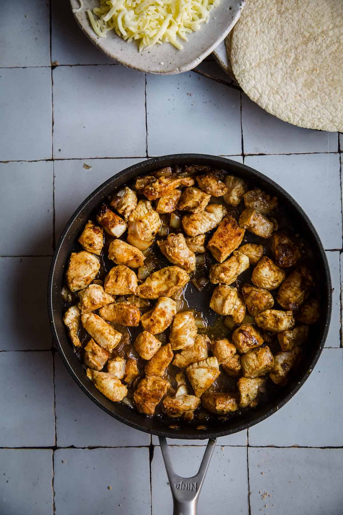 Chicken pieces in a black frying pan on a tiled background.