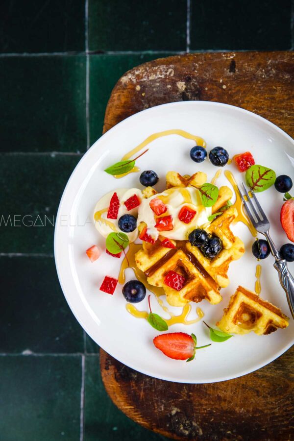 a waffle and fruit on a white plate with green tiles in background.