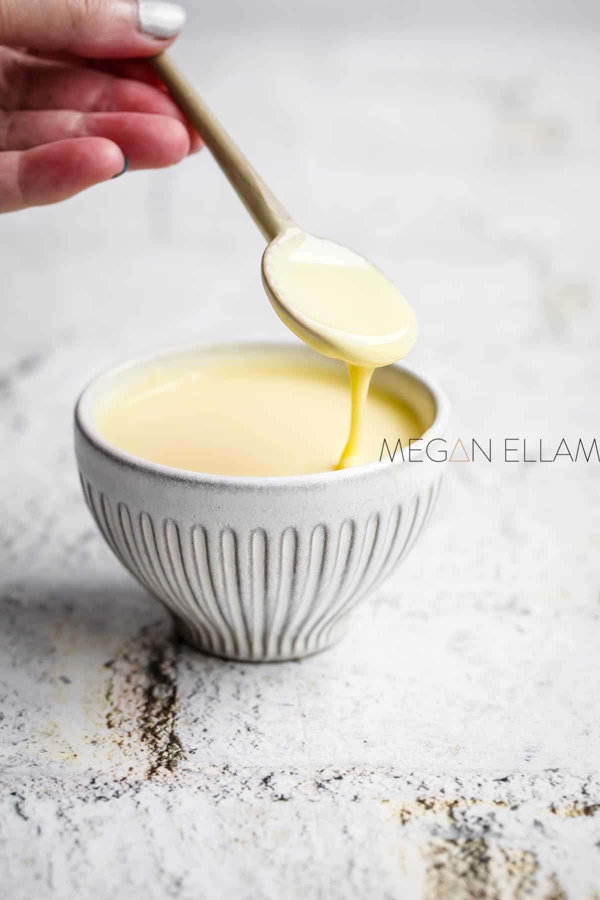 A womans hand holding a spoon of sugar free condensed milk above a bowl of it.