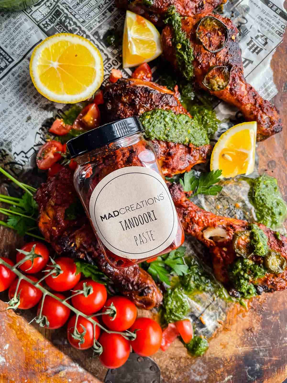 A jar of homemade tandoori pastes on top of baked pieces of chicken, tomatoes and herbs.