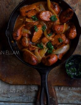 Curried Sausages in a cast iron pan on a wood chopping board.